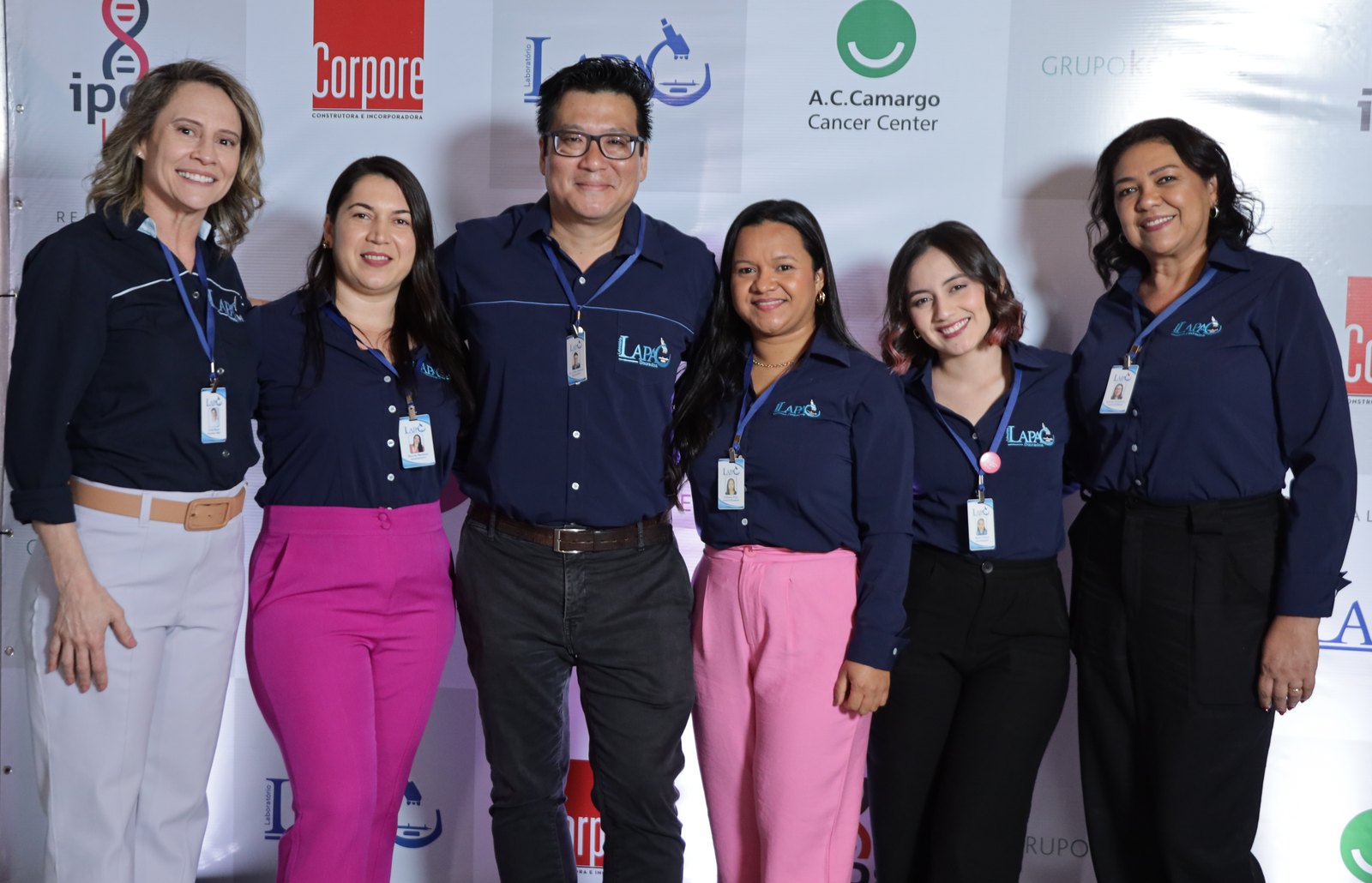 Equipe do LAPAC, de camisa azul-marinho com o logotipo do laboratório, em frente ao painel do simpósio