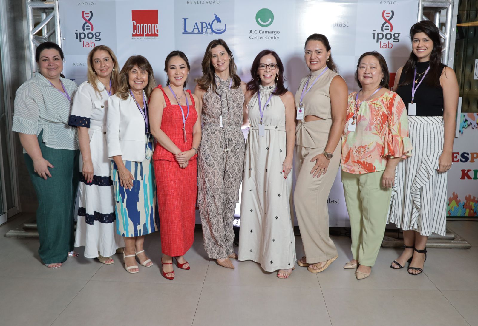 Nove participantes com crachá do simpósio em frente ao painel dos realizadores e apoiadores